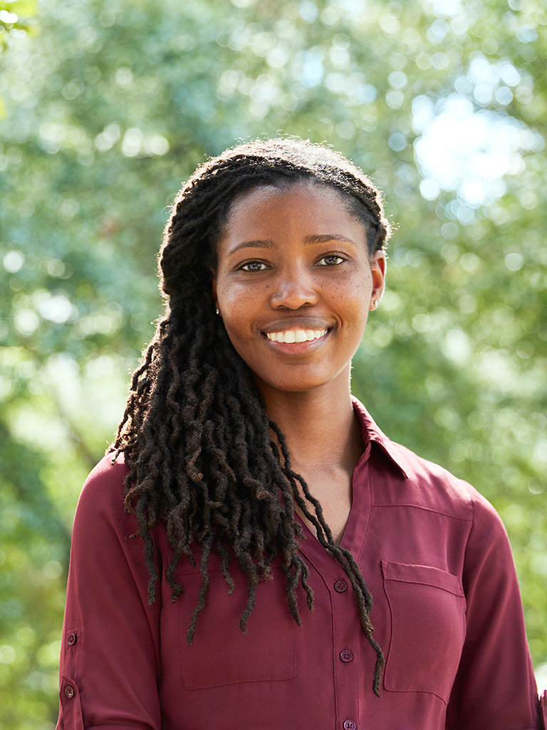 A woman stands, smiling at the viewer. A background of bright, green foliage is behind her.